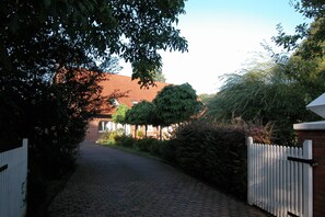 A paved pathway leading to a building with a red roof, surrounded by greenery and a white picket fence. - Vacation apartment at Wulfkamp (Velen)