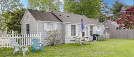 A single-story house with a white exterior, a front porch with two Adirondack chairs, a picnic table, and a barbecue grill.