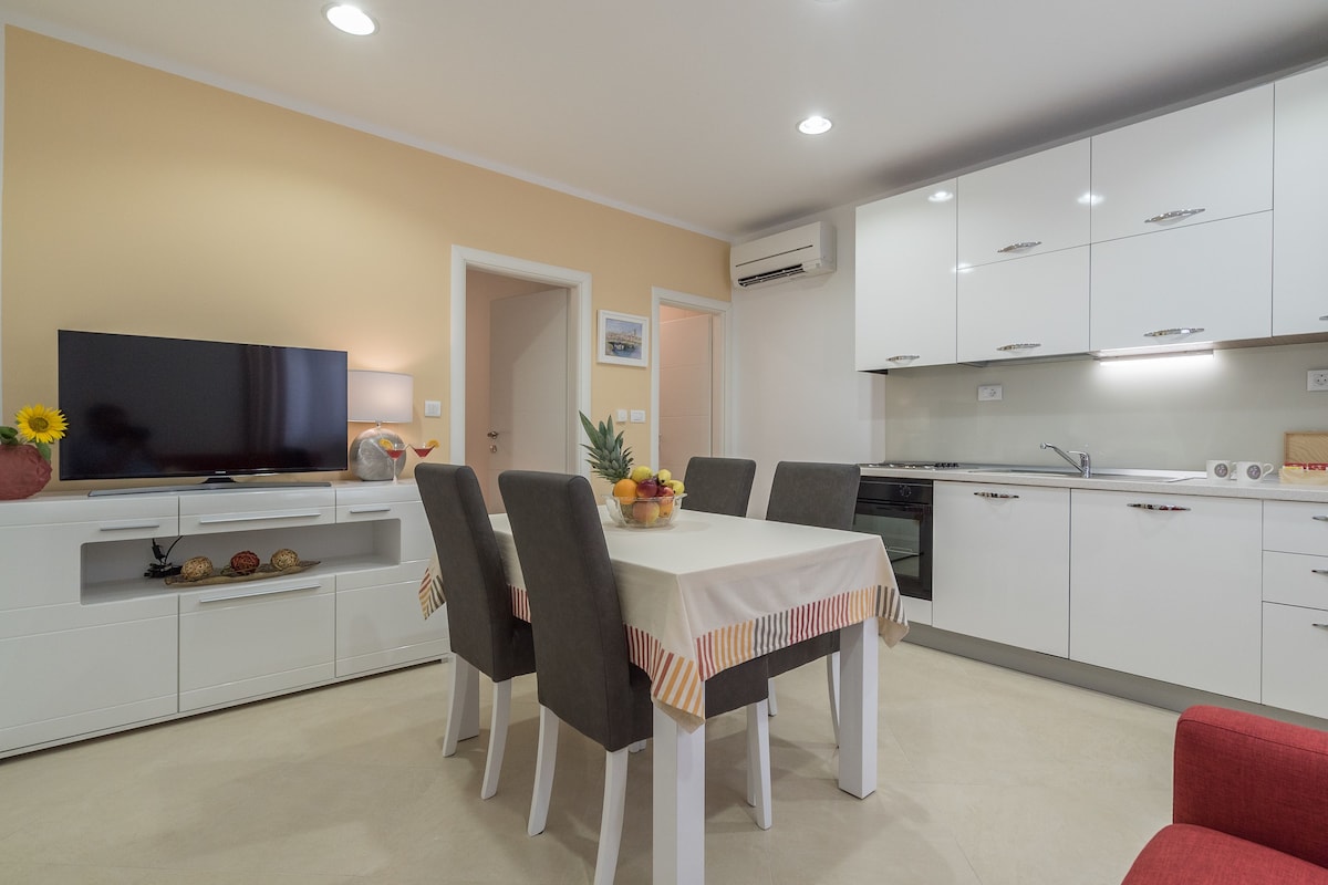 A modern kitchen with white cabinets, a dining table with chairs, a TV, and a fruit bowl.