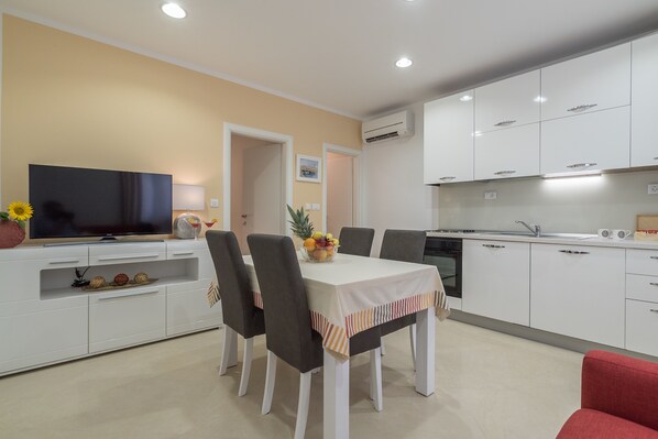 A modern kitchen with white cabinets, a dining table with chairs, a TV, and a fruit bowl. - Drop In Split Suites (Split)