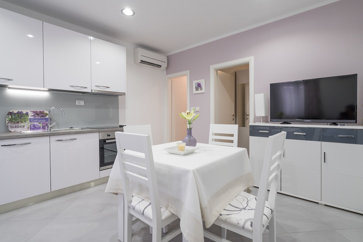 A modern kitchen with white cabinets, a dining table with white chairs, a TV, and a vase with flowers.