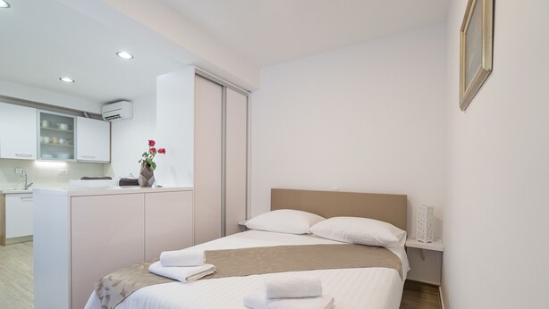 Habitació d'hotel moderna amb un llit ben fet, una petita taula de nit amb una làmpada i vista a la zona de la cuina.