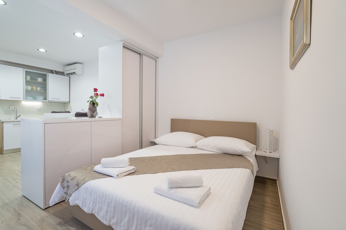 A modern hotel room with a neatly made bed, a small bedside table with a lamp, and a view of the kitchen area.