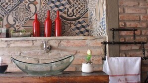 Casa de banho com lavatório em vidro, garrafas vermelhas, uma planta em vaso e um porta-toalhas com toalha.