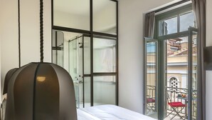 Quarto com cama, mesa de cabeceira, luminária pendente, janela com cortinas e vista para uma varanda externa.