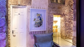 Quarto com arco de pedra, porta branca, obra de arte emoldurada, poltrona de couro e pequena mesa com um livro.