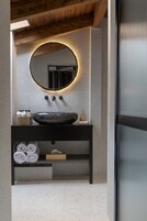 A modern bathroom with a large round mirror, a black vessel sink, and neatly rolled towels on a shelf.
