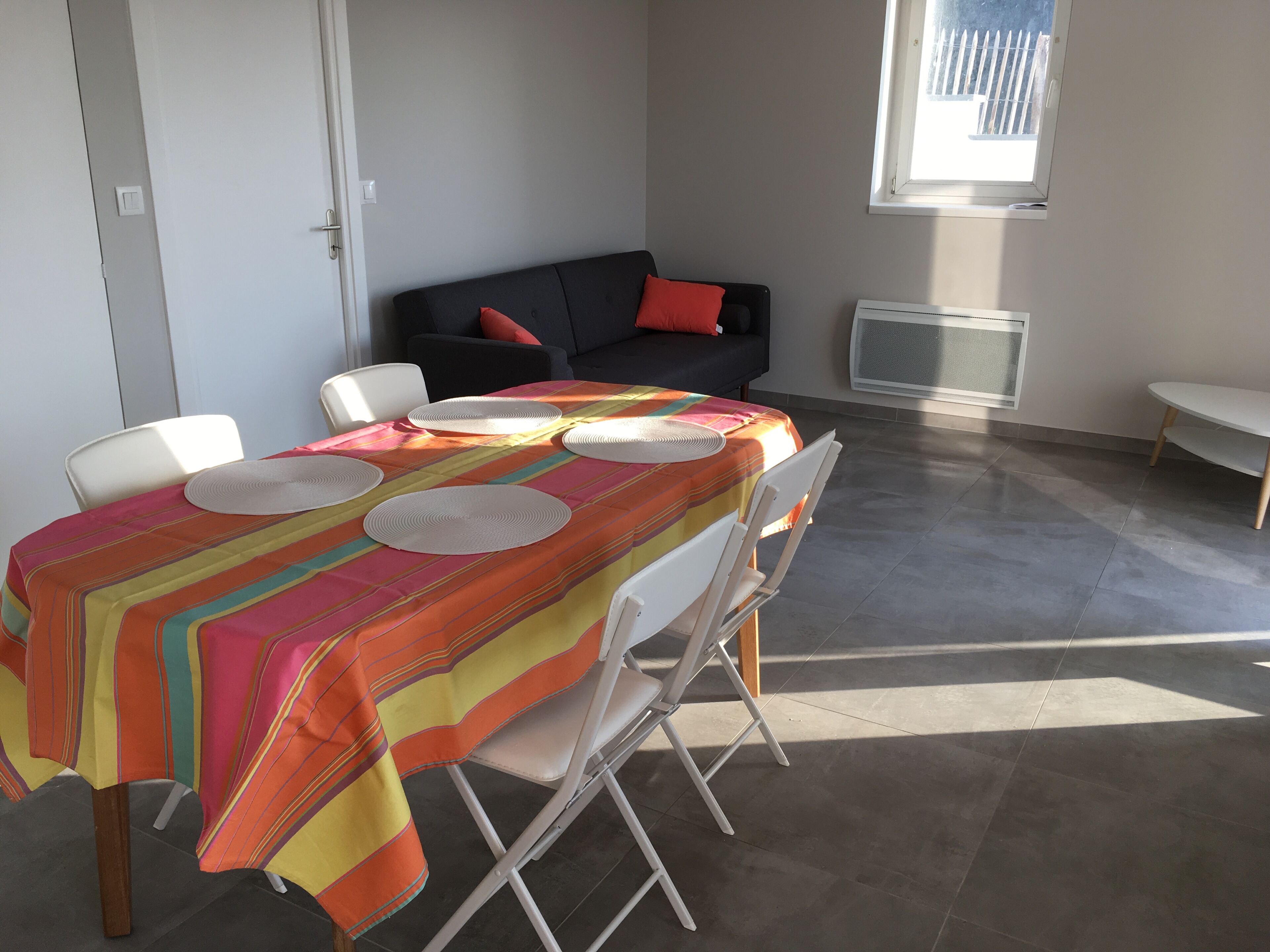 Un coin repas avec une table dressée pour quatre personnes, des chaises blanches et une nappe rayée aux couleurs vives.