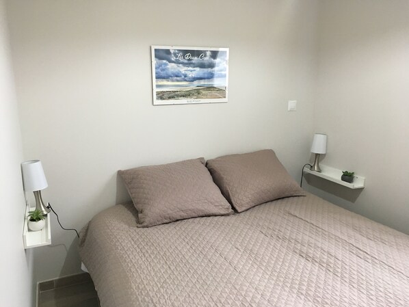 A bedroom with a bed, two bedside lamps, a small shelf with a plant, and a framed picture on the wall.