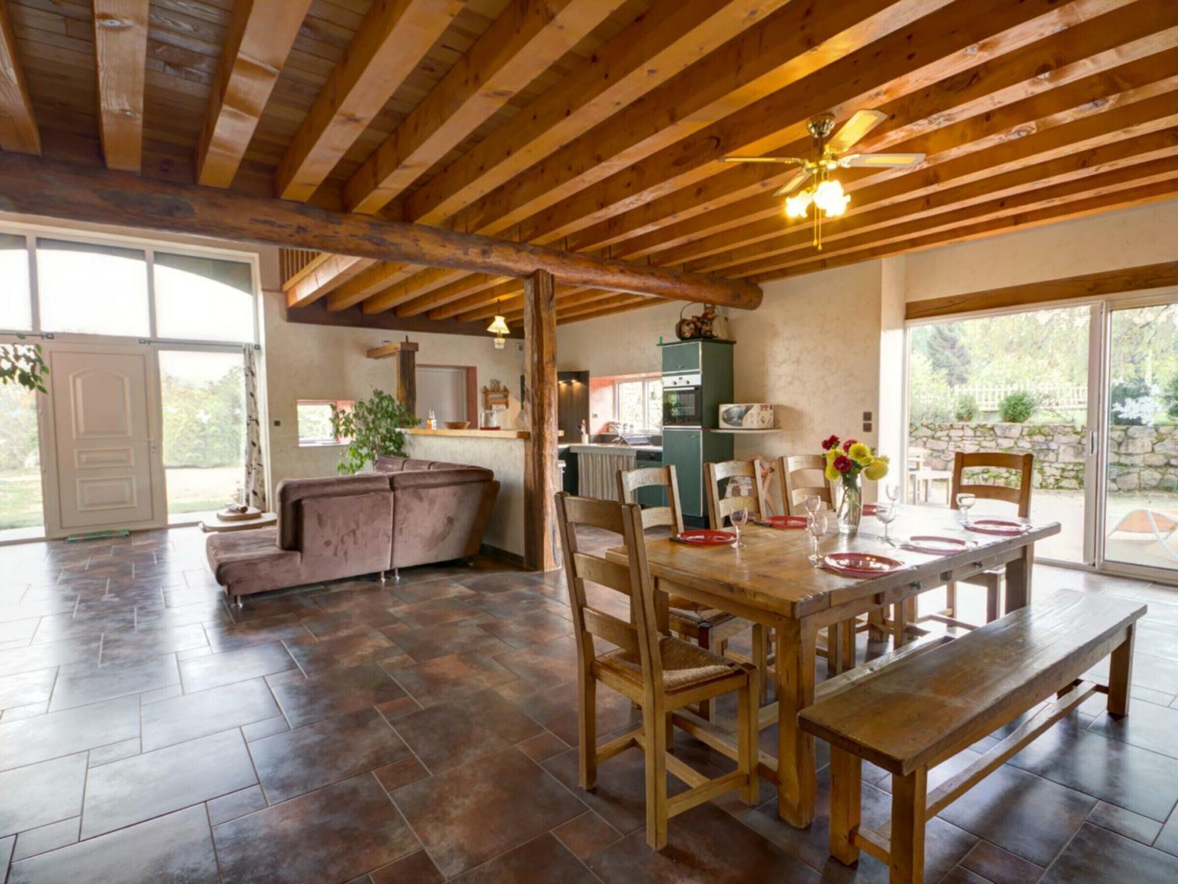 Un comedor amplio con una mesa y bancos de madera, una cocina al fondo y un gran ventanal corredera que da a un patio exterior.