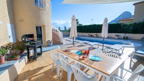 Une terrasse extérieure avec une table à manger pour quatre personnes, entourée de chaises en plastique blanches. Un barbecue est visible sur la gauche, et une piscine se trouve au fond.