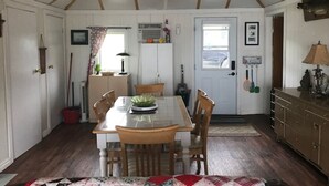 A dining area with a wooden table and chairs, a kitchen with a refrigerator and a sink, and a door leading outside. - PEACEFUL OASIS COTTAGE (Keene)