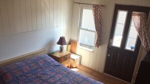 A bedroom with a bed, a nightstand, a lamp, a window with curtains, and a door. - PEACEFUL OASIS COTTAGE (Keene)