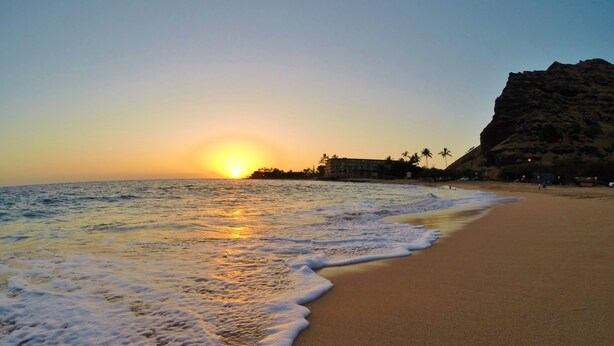 A beach with a sunset, waves, and a rocky cliff.