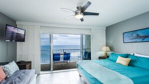 A bedroom with a bed, a TV, a ceiling fan, and a balcony with a view of the ocean.