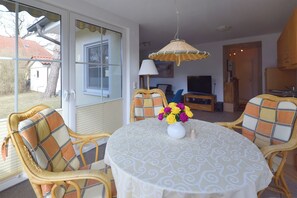 A dining area with wicker chairs, a round table with a patterned tablecloth, a hanging light fixture, and a window with a view of a house. - Centrally located apartment for 4 people, south-west facing terrace, washing machine, WLAN (Ostseebad Prerow)