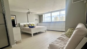 A modern bedroom with a bed, a white sofa, a ceiling fan, and a large window with a city view. - Beach front, best location, ocean and city views + parking! (San Juan)