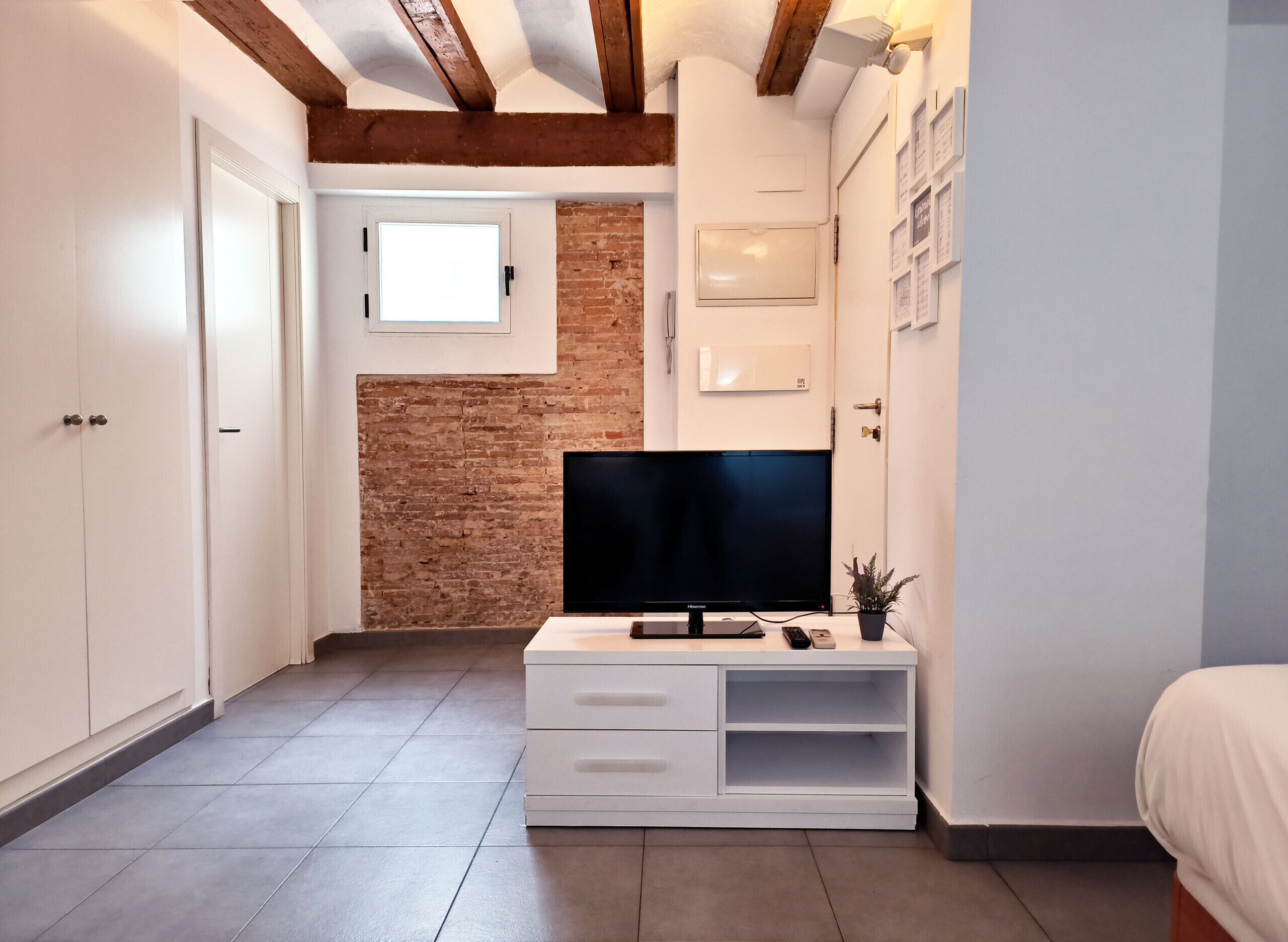 A small, modern room with a TV on a white TV stand, a brick wall, and a window.