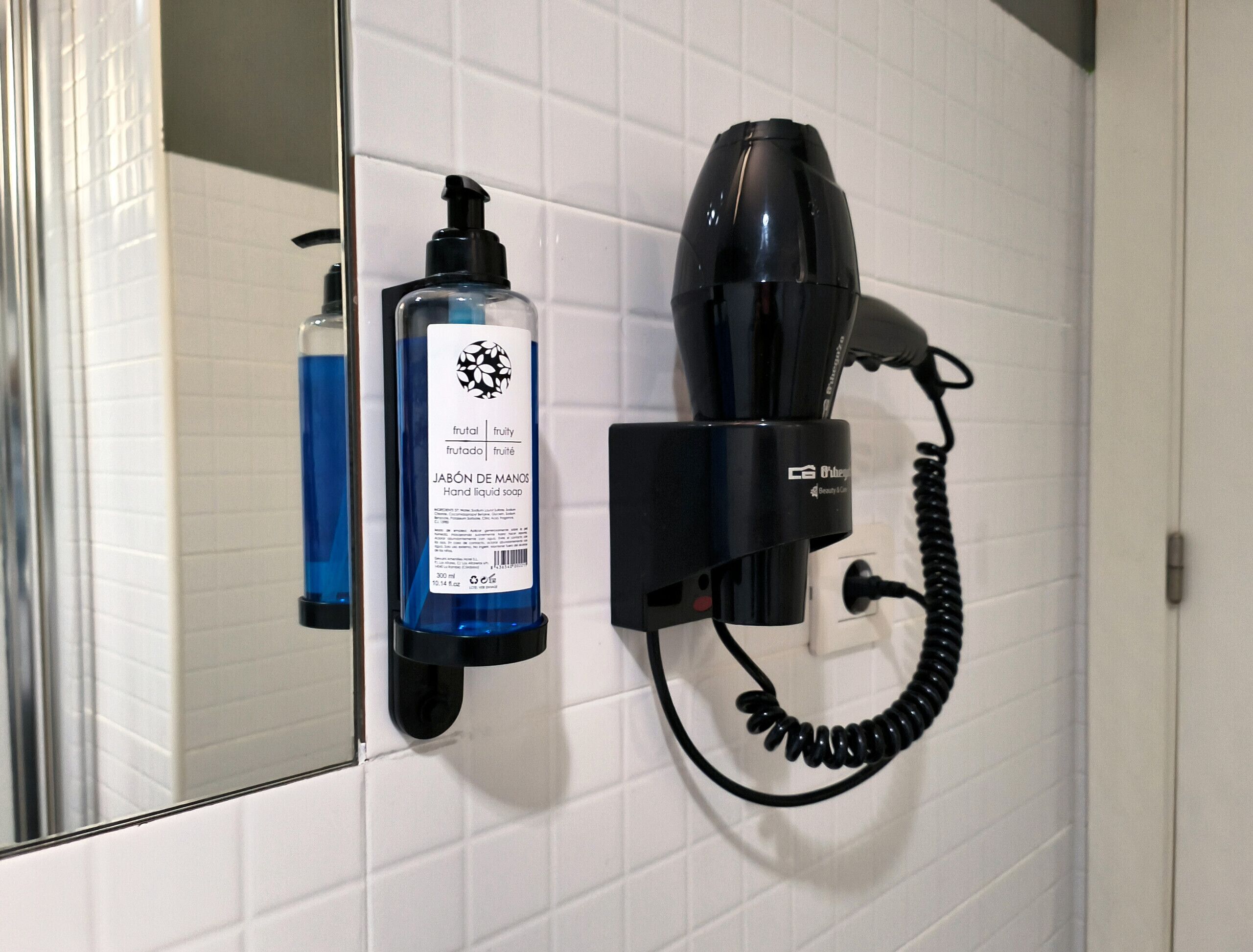 Un dispensador de jabón para manos y un secador de pelo fijados a una pared de azulejos blancos.