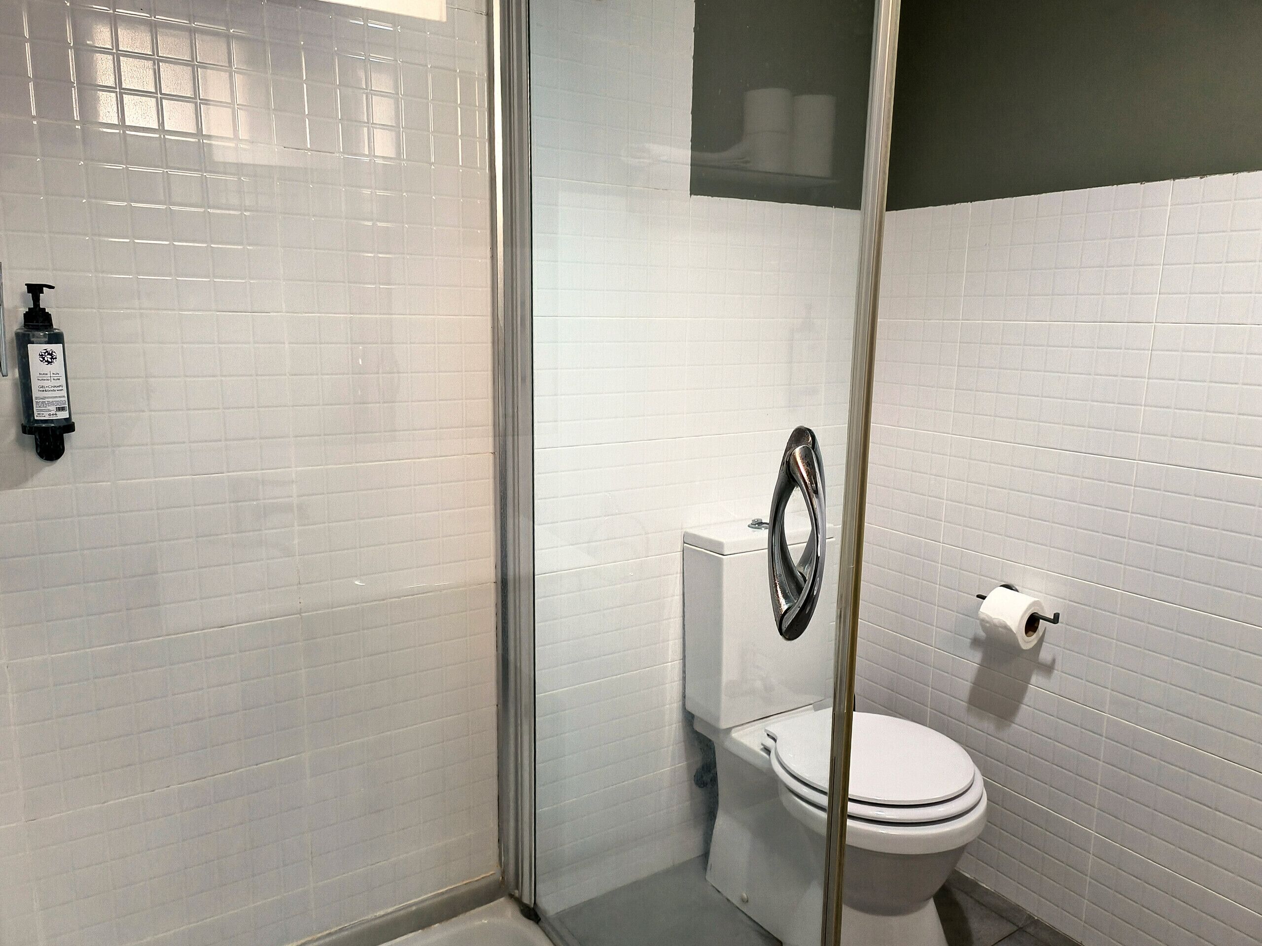 Ein Badezimmer mit weißen Fliesen, einer Glasdusche, einer Toilette und einem an der Wand montierten Seifenspender.