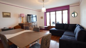 A living room with a sofa, a dining table with chairs, a TV on a cabinet, and a large window with a view. - Spacious Apartment in Evian with Lake View, Balcony, and Pet-Friendly (Évian-les-Bains)