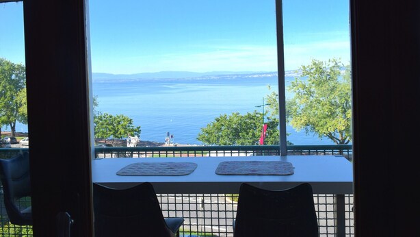 A table with two placemats, a chair, and a view of a body of water with trees and a distant shoreline. - Spacious Apartment in Evian with Lake View, Balcony, and Pet-Friendly (Évian-les-Bains)