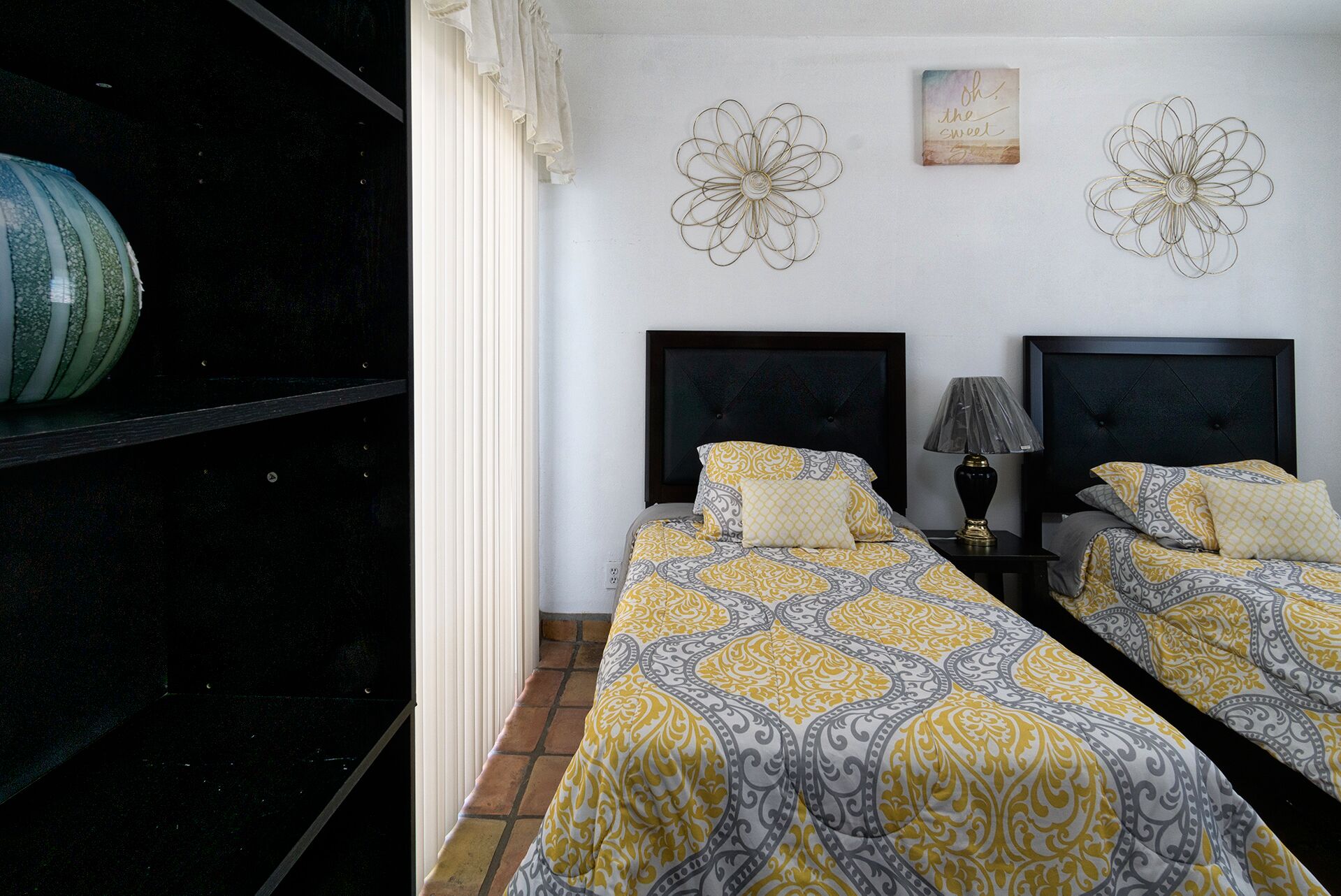 Habitación de hotel con dos camas, cada una con colcha estampada en amarillo y gris. Se observan obras de arte decorativas sobre las camas y, a la izquierda, una estantería con un jarrón verde.
