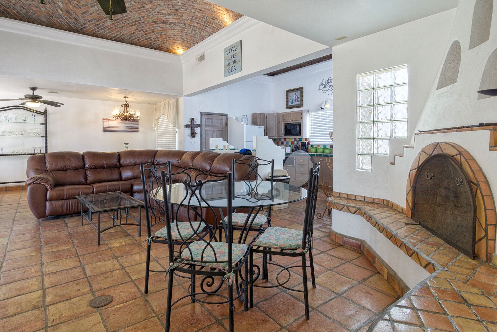 A spacious living area with a brown leather sofa, a glass-top dining table with chairs, and a kitchen in the background.