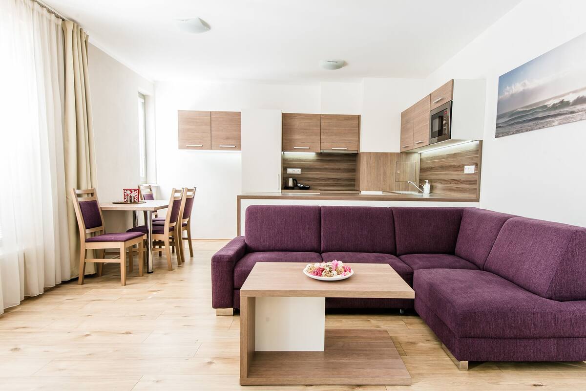 A modern living room with a purple sofa, wooden dining table, and a kitchen area with wooden cabinets and a built-in oven.