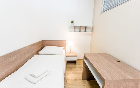 A small, neatly arranged bedroom with a wooden bed, a bedside table with a lamp, a small shelf with a plant, and a bench. - Apart Hotel VIRGO (Bratislava)