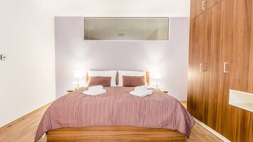 Una cama bien hecha con una colcha rosa, dos almohadas blancas y una toalla blanca doblada al lado. A la derecha hay un armario de madera y una ventana con persianas sobre la cama.