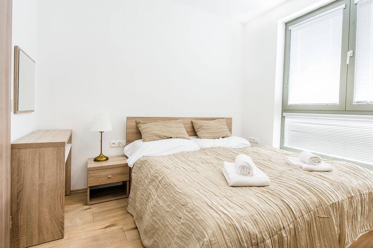 A bedroom with a wooden bed, a nightstand, a lamp, and a window with blinds.