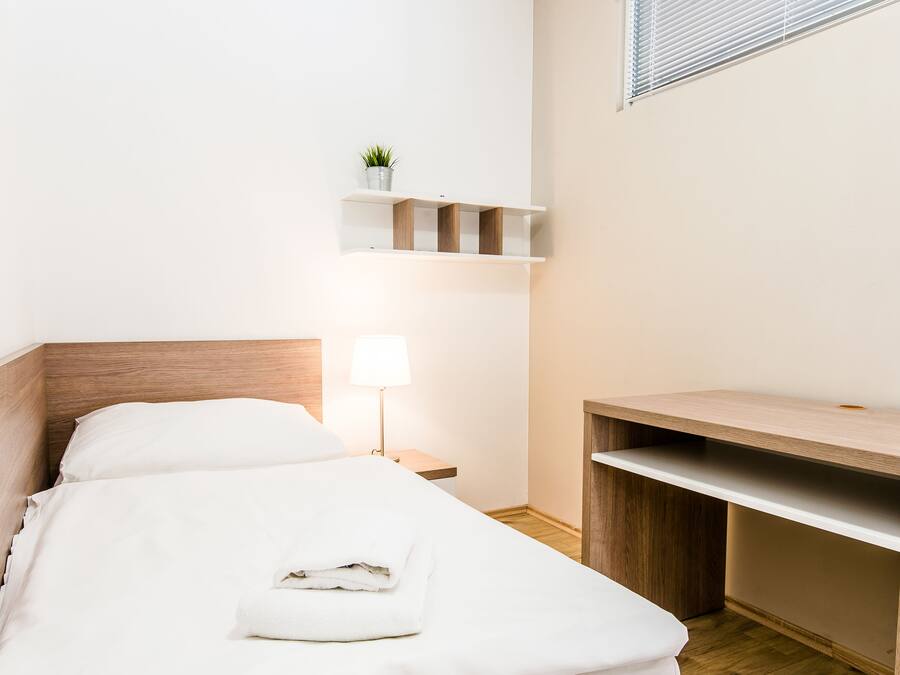 A neatly made bed with white linens, a bedside lamp, a wooden nightstand, and a small shelf with a plant.