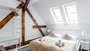 Een slaapkamer met een schuine plafond, houten balken, een bed met witte lakens, twee nachtkastjes en een raam.