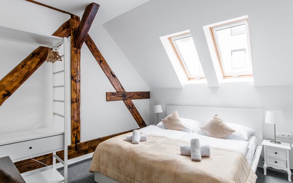 Un dormitorio con techo inclinado, vigas de madera, una cama con ropa blanca, dos mesitas de noche y una ventana.