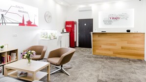 Een receptiegedeelte met een houten bureau, twee bruine stoelen, een rode Coca-Cola koelkast, een wereldkaart en een bord met begroetingen in meerdere talen.