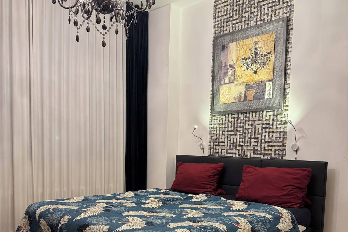 Un dormitorio con una cama grande, una araña, un papel tapiz decorado y una ventana con cortinas.