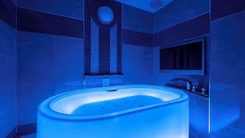 A modern bathroom with a large, illuminated bathtub, a round mirror, and a sleek, dark-toned sink.