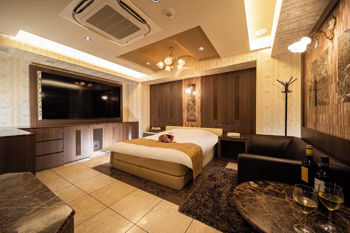 A modern hotel room with a large bed, a flat-screen TV, a minibar, and a seating area with two wine glasses.