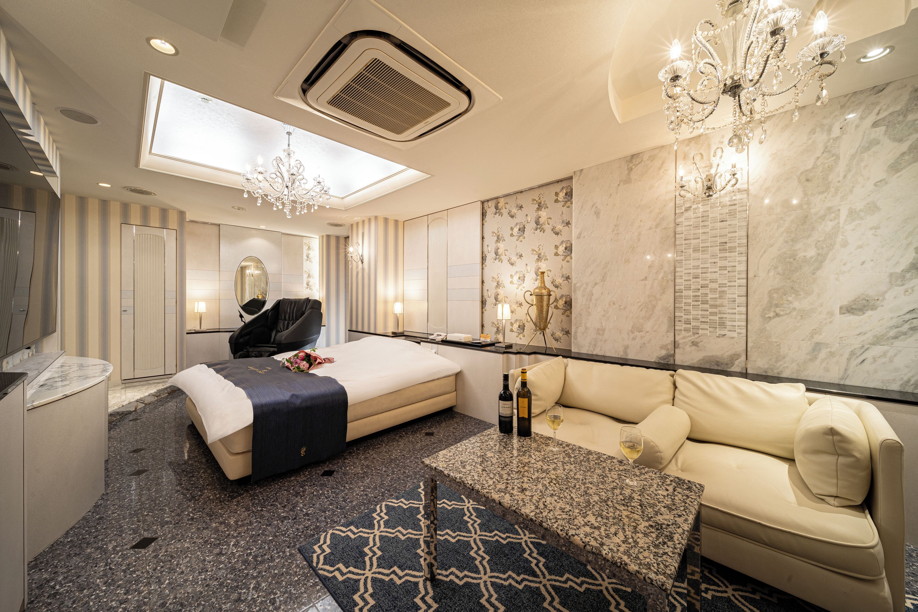 Quarto de hotel moderno com uma cama grande, um sofá, uma pequena mesa com garrafas e um candelabro.