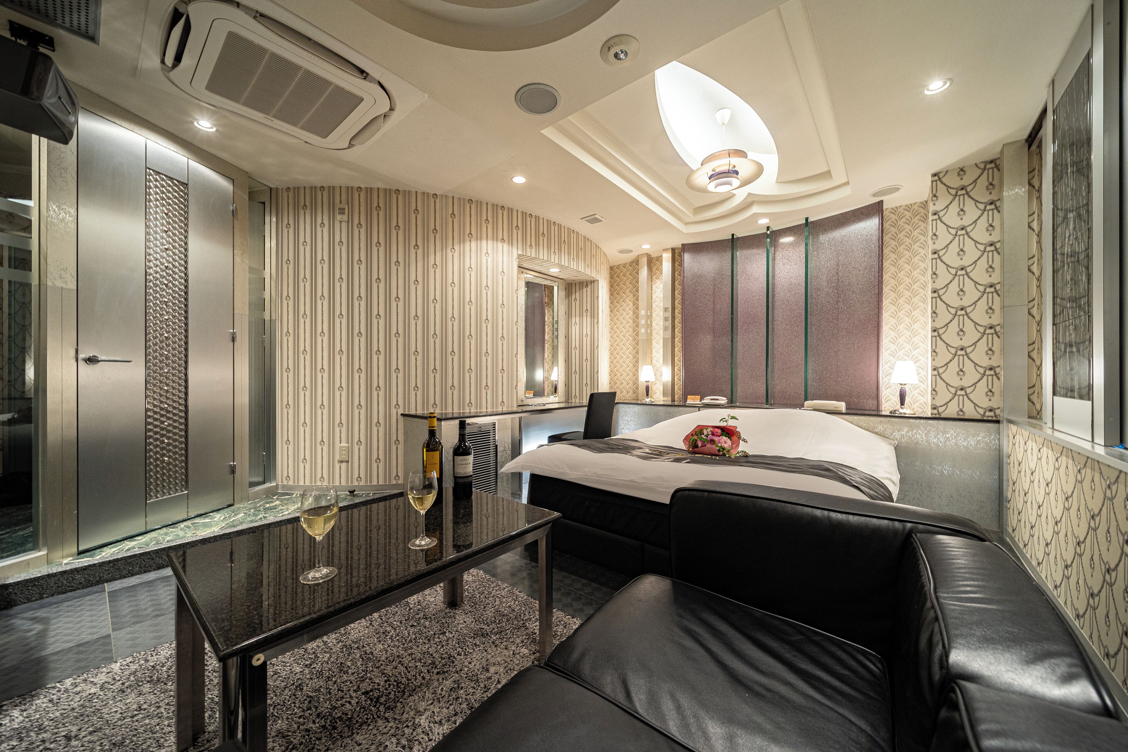 Une chambre d’hôtel moderne avec un lit, une table en verre sur laquelle se trouve une bouteille de vin, un canapé en cuir et un grand miroir.