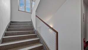 Una escalera con escalones de granito y barandilla de metal, que conduce a un rellano con una ventana y un extintor en el suelo.