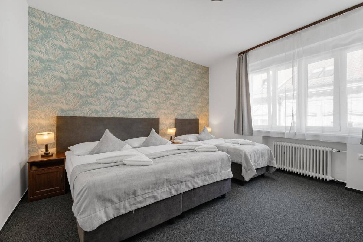 Habitación de hotel con dos camas, un radiador, una ventana con cortinas traslúcidas y un papel tapiz estampado.