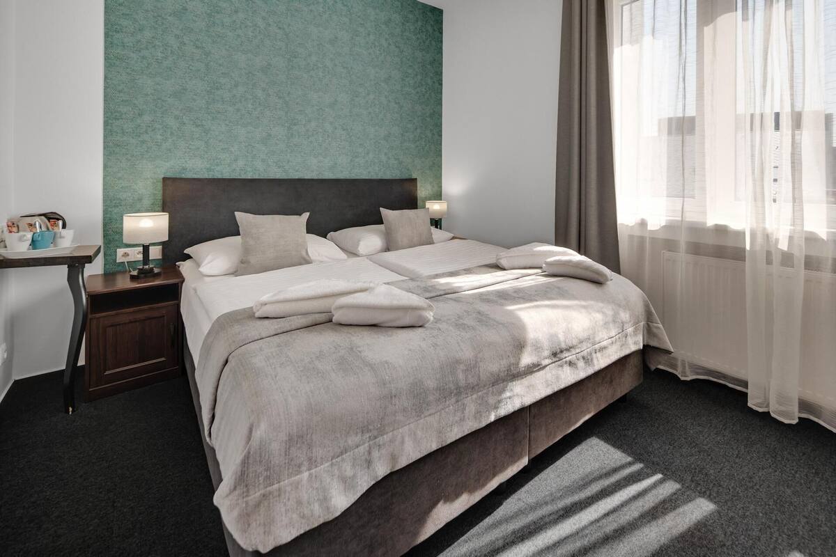 Habitación de hotel con una cama, mesitas de noche, una lámpara y vistas a la ciudad a través de cortinas translúcidas.