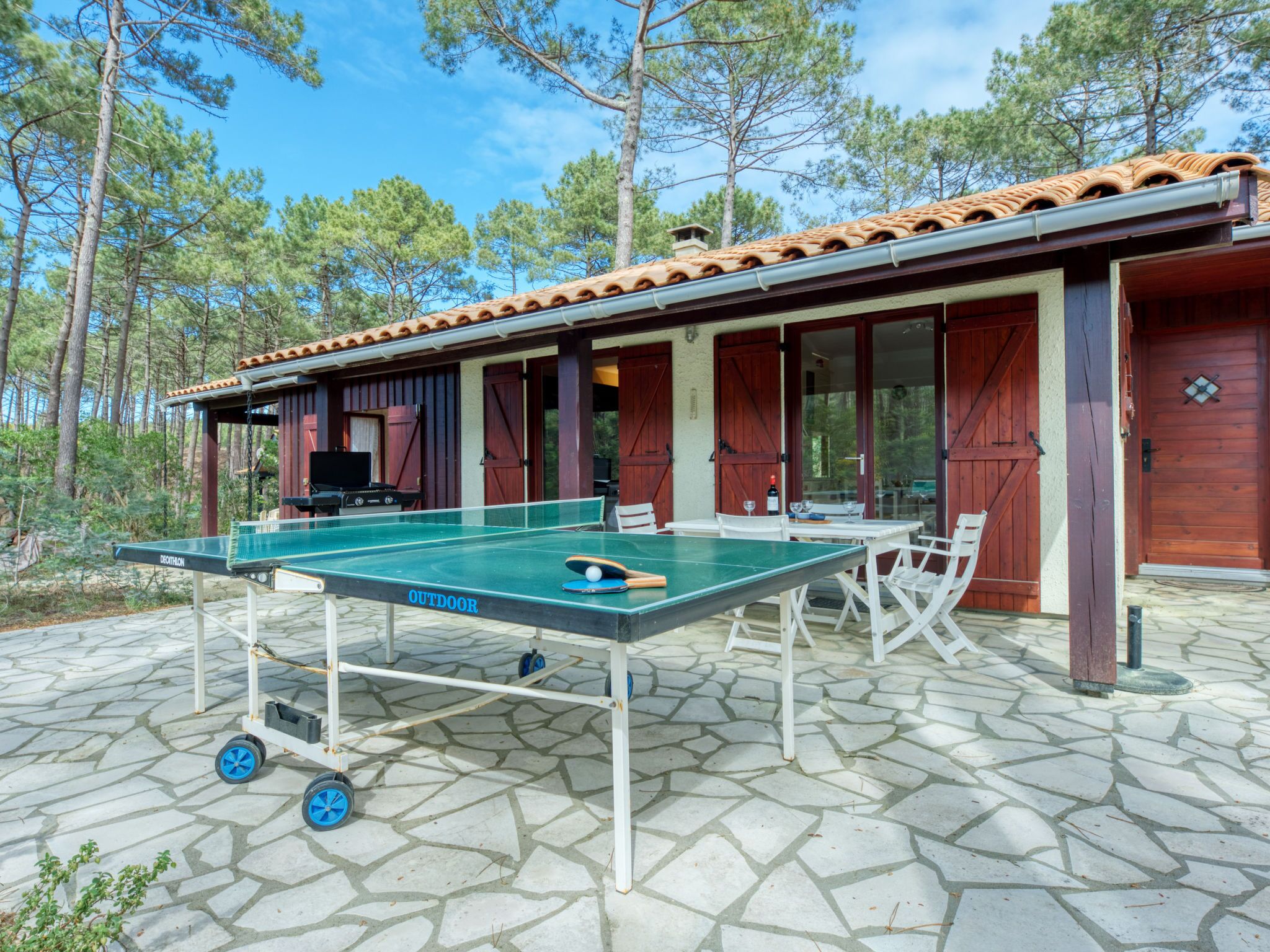 Une table de ping-pong installée à l’extérieur, sur une terrasse en pierre, avec un bâtiment en bois en arrière-plan.