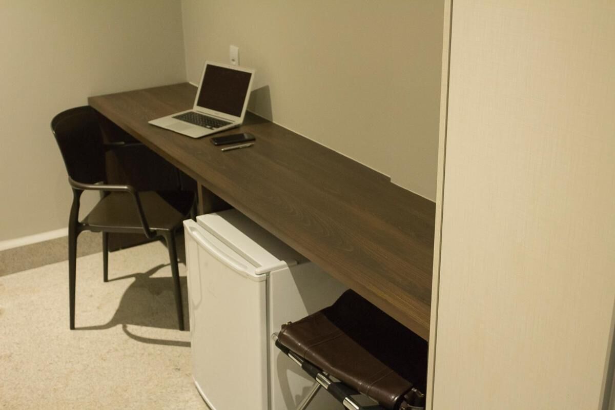 Un espacio de trabajo reducido con una computadora portátil, un teléfono inteligente, una silla y un refrigerador pequeño.