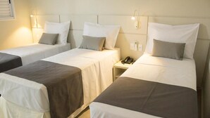 A hotel room with three single beds, each with a white and gray duvet, a bedside table with a phone, and a wall-mounted light fixture.