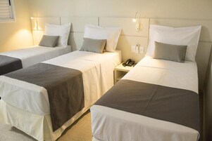 Quarto de hotel com três camas de solteiro, cada uma com colcha branca e cinza, mesa de cabeceira com telefone e luminária fixada na parede.