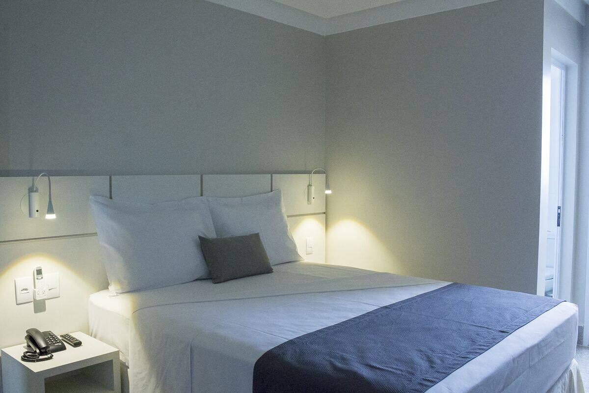 A neatly made bed with white linens and a dark blue accent, a bedside table with a phone and remote, and a wall-mounted light fixture.