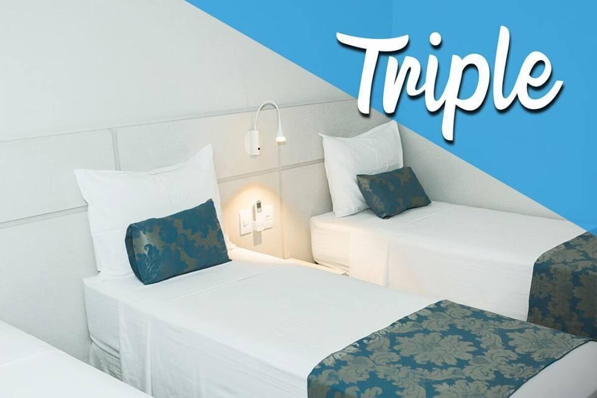 A hotel room with three single beds, each with a white headboard and a blue patterned bedspread. There are bedside lamps and a wall-mounted light fixture.
